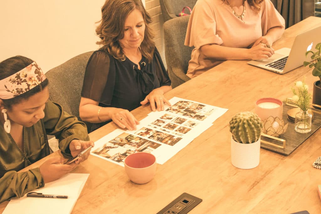 Three women sit at a long table, looking at mood boards while talking