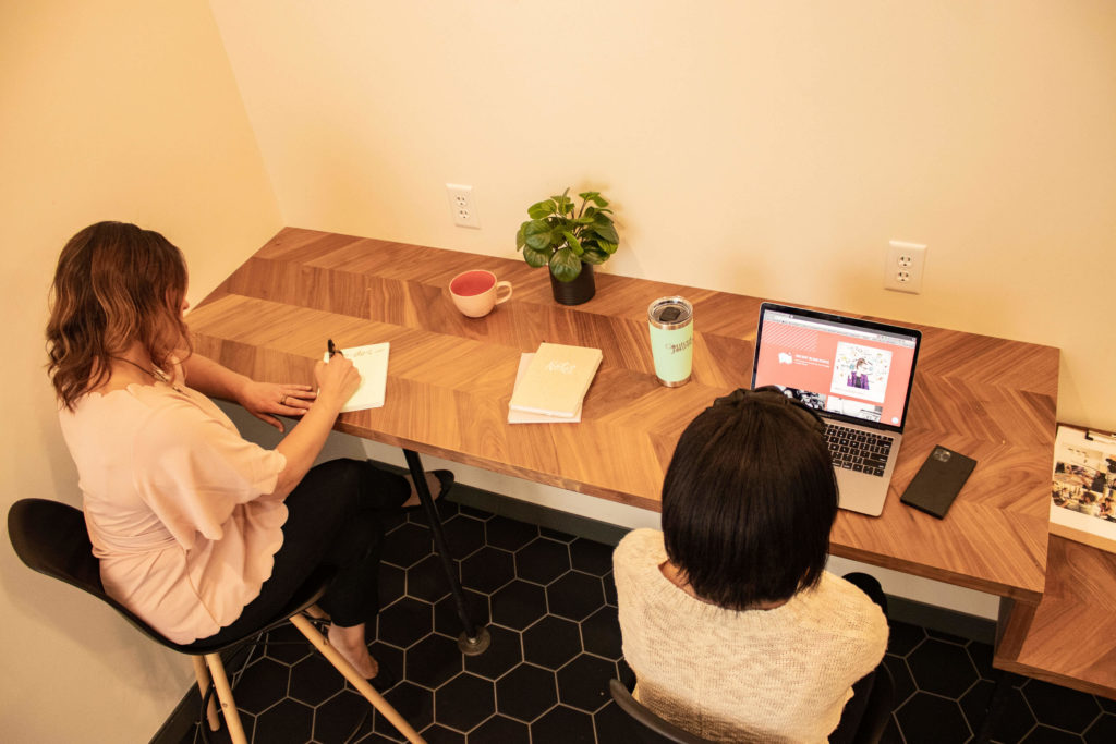 Two women sit at a long table against a wall. One is writing in a notebook the other is on a computer. Their backs are to the camera