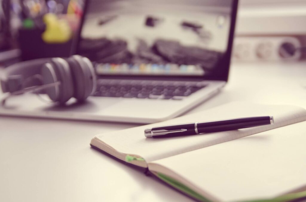 A pen laying on an open notebook with a computer and headphones in the background
