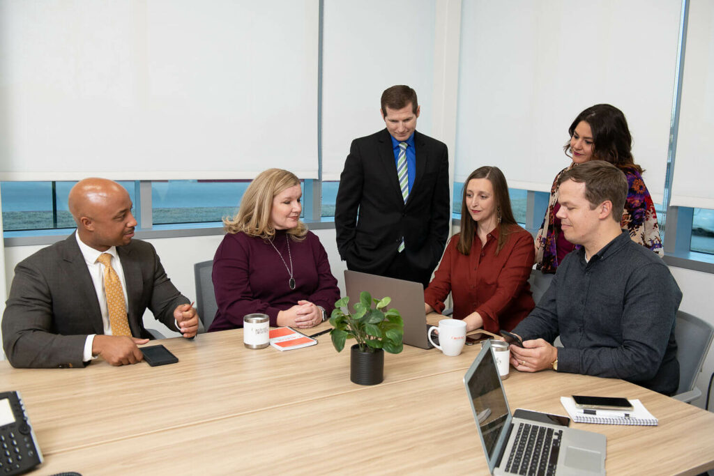 A group of people around a conference room table looking at a laptop together.