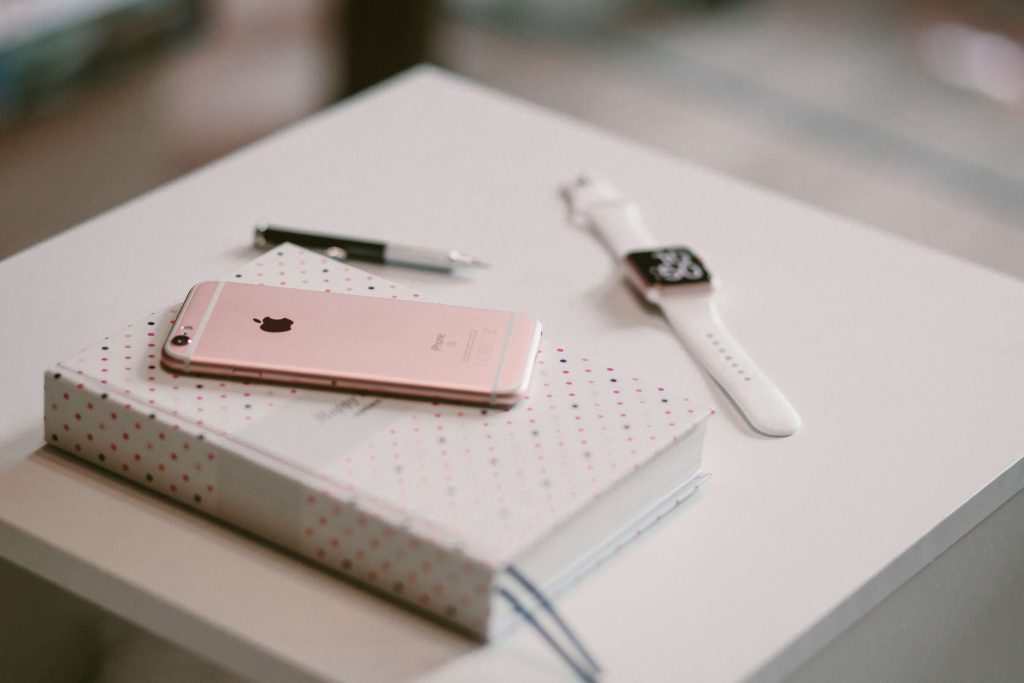 A journal with a pink eye phone on top of it and an Apple watch laying off to the side on a white table